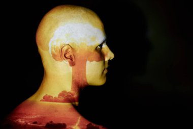 Profile of emotionless bald man with image of cloud from nuclear explosion projected on face standing against black background in studio, copy space clipart