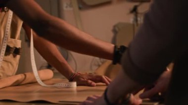 Önlük giymiş genç bir Afro-Amerikan zanaatkar, deri detaylarını ölçerken, ekipmanlı ve sıcak ışıklandırmalı bir atölyede tanınmayan meslektaşıyla birlikte.