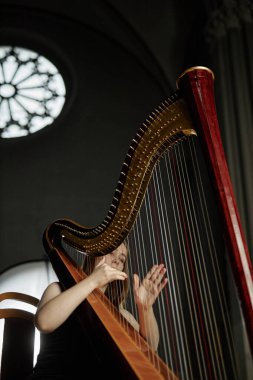 Antik ahşap arp ile müzik çalan kadın müzisyenin dikey çekimi. Kilisede loş ışıkla çalarken hafifçe tellere dokunuyor. Kopya alanı.