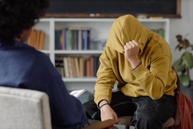Stresli genç çocuğun portresi terapistle ruh sağlığı sorunlarını paylaşırken bir seansta sandalyeye oturup yüzünü gizlemesi.
