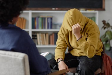 Portrait of distressed teenage boy sharing mental health troubles with therapist while sitting on chair and hiding face during one on one session copy space clipart