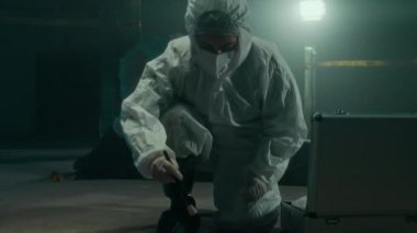 Kadın adli tıp uzmanının, iş arkadaşıyla birlikte, arka planda olay yerinde çalışan plastik torba altında cesedi incelemek suretiyle kanıt toplaması.