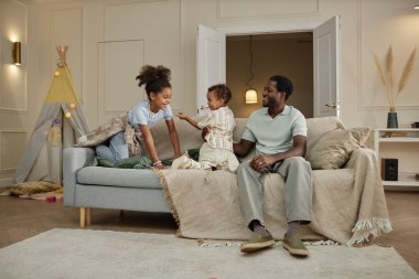 Gülümseyen Afrikalı Amerikalı çocukların babalarıyla kanepede oynamalarının geniş açı çekimi. Hafta sonları sıcak oturma odasında birlikte eğlenmeleri, mutlu aile sahneleri.