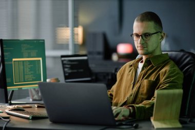 Uzun iş gününün sonunda bilişim şirketinde masasında otururken dizüstü bilgisayarda kod yazan kendinden emin bir erkek bilgisayar programcısının orta boy görüntüsü.