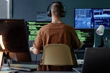 Ofiste tek başına çalışırken birden fazla bilgisayarda yazma koduna boğulmuş kulaklık takan erkek bilişim geliştiricisinin arka görüntüsü