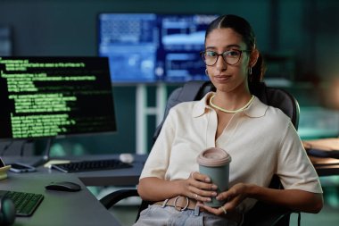 Orta boy Orta Doğulu pozitif kadın bilişim geliştiricisinin iş yerinde elinde kahve ile iskemlede otururken çekilmiş portresi.