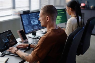 Modern teknoloji şirketinde çalışma masasında çalışırken dizüstü bilgisayara gömülü minimalist küpeli şık erkek bilişim programcısının yan görüntüsü.