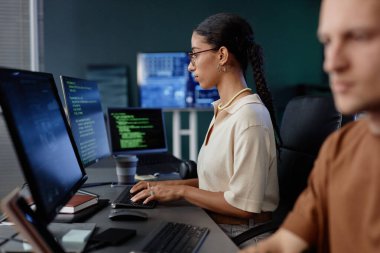 Profesyonel Ortadoğulu kadın programcının yan görüntüsü bilgisayardaki yazım kodlarına yoğunlaşırken aynı zamanda bilişim şirketinin ofisindeki iş yerinde meslektaşlarıyla oturuyordu.