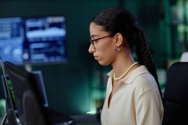 Bilişim şirketinin ofisindeki bilgisayardaki program için zarif gümüş küpeler ve gözlük takan Orta Doğulu kadın müteahhitin yan görünüm portresi.