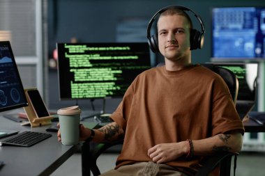 BT şirketinin ofisinde çalışırken kameraya güvenle bakan ve elinde bir fincan kahveyle oturan, kulaklık takan pozitif erkek programcının orta boy portresi.