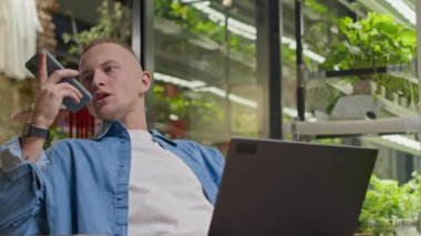 Genç, kendine güvenen beyaz bir adamın laptopun önündeki koltukta otururken ve arka planda bulanık yeşil dikey çiftliğin cam duvarlarıyla telefonda sesli mesaj kaydettiği orta boy bir fotoğraf.