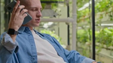 Gündelik giyimli genç beyaz adamın laptopta yazarken birden fazla iş yapan ses kaydını çekerken, yeşil dikey çiftliğin cam duvarlarının önünde otururken.