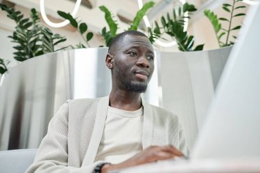 Portrait shot of African American businessman looking at laptop screen working in lounge zone of contemporary coworking space with biophilic interior design, copy space clipart