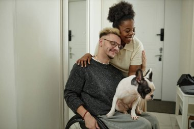 Orta boy, neşeli gülüşü olan Afrikalı Amerikalı bir kadının sevgilisine sevgiyle sarılırken tekerlekli sandalyeyi bir çift olarak kullanırken evcil köpek eşliğinde, mutlu bir ilişki sahnesi paylaşırken.