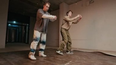 Sokak kıyafetleri içinde iki yetenekli beyaz erkek zoomer 'ın enerji ve stil dolu yaratıcı kareografik break dansı yaparken, kapalı alanda pratik yaparken, elde çekilmiş bir fotoğrafı.