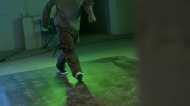 Şehir kıyafetleri içinde sakallı genç beyaz çocuk yeşil ve mavi ışık altında kapalı alanda dans ederken rock 'n' roll hareketleri sergiliyor.