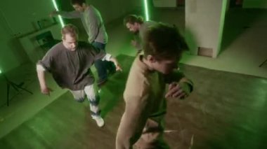 Üç beyaz erkek zoomerın yüksek açılı çekimi ve dans eden hip hop yapan genç adam dans pistinde yeşil neon ışıklarıyla aydınlatılarak daireler çiziyorlar.