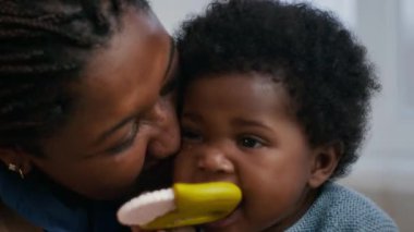 Gülümseyen Afrikalı Amerikalı annenin bebeğini öperken ve kucaklarken, diş çıkarma oyuncağını kemirirken yakın plan fotoğrafı.