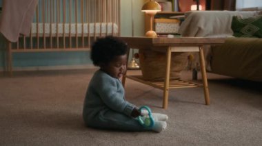 Mavi elbiseli Afro-Amerikalı çocuk çıngırak diş çıkarma yüzüğüyle oynuyor, halı döşeli yatak odasında oturuyor.