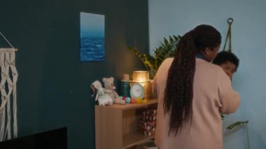 Uzun örgülü Afro-Amerikan annesi bebeğini kucağında beşikte rahat bir bebek odasında bir sürü oyuncakla ve duvar süslemeleriyle uyuyor.