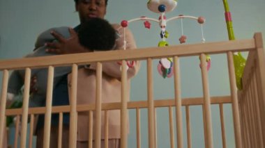 Afrika kökenli Amerikalı annenin ahşap karyolaya eğildiği, kıvırcık saçlı bebeği rahat bir bebek odasına renkli bir cep telefonunun altına yerleştirdiği düşük açılı fotoğraf.