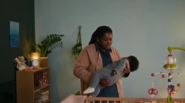 Afrika kökenli Amerikalı anne, renkli mobil süslemeli beşikteki beşikteki bebeğini dikkatlice yerleştiriyor.