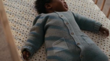 Sevimli Afro-Amerikan bebek mavi örülü tulum giyiyor, altın yıldızlı beyaz çarşafla süslenmiş ahşap beşiğinde kestiriyor.