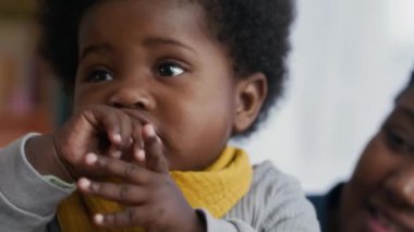 Sarı önlüklü şirin Afro-Amerikan bebek annesinin kucağında gevezelik ederken yakın plan çekimleri.