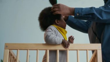 Düşük açılı, tanınmayan Afrikalı Amerikalı annenin, bebeğin kıvırcık saçlarını tararken, ahşap karyolada dururken.