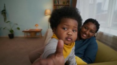 Annesi onu kucağına alıp gülümserken, sarı önlüklü Afrikalı Amerikalı çocuğun koltuktan kamera almaya çalışmasının portresi.