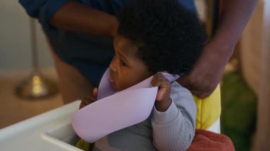 Afro-Amerikalı çocuk huysuz ve önlük giymeyi reddediyor, öğle yemeği sırasında yüksek sandalyede oturuyor.