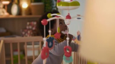 Afro-Amerikalı bebek ayakta duruyor, taşınabilir süslemeli beşiğe yaslanıyor ve rahat çocuk odasında U şeklinde diş fırçasını kemiriyor.