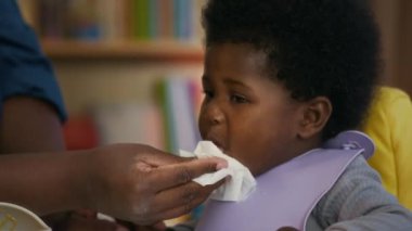 Onu yüksek sandalyede besledikten sonra çocuğun yüzünü ve ellerini silen tanınmamış Afrikalı Amerikalı annenin yakın plan fotoğrafı.