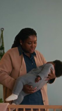 Afrikalı Amerikalı anneyi sevmenin dikey görüntüsü bebeğini beşiğine koyuyor ve onu rahat ettirmeye çalışıyor.