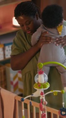 Dikey çekimde Afrikalı Amerikalı anne bebeğini ahşap bir beşikte rahat bir çocuk odasına yerleştiriyor.