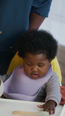 Dikey çekimde, Afro-Amerikan annesinin aç ve heyecanlı yürümeye başlayan çocuğuna yemek kasesi göstermesi. Yüksek sandalyede oturuyor.