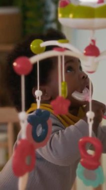 Dikey çekimde, Afrikalı Amerikalı bebek gevezelik ediyor ve etrafa bakıyor, beşiğinde duruyor ve U şeklindeki diş fırçasını kemiriyor.