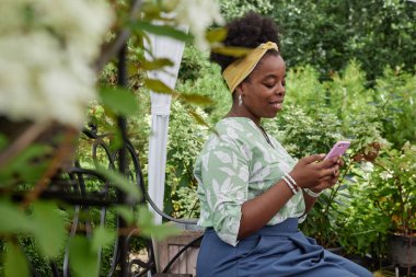 Orta boy Afro-Amerikan kadın peyzaj tasarımcısı müşterilerle iletişim kurarken bahçe merkezinde canlı yaz yeşilliğinin keyfini çıkarırken cep telefonuna mesaj yazıyor.