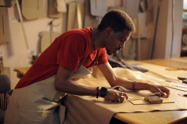 Zanaatkar dikkatle dikkatle dikkatle deri parçaları üzerinde çalışıyor. Ahşap bir masada deri keserken ve şekillendirirken önlük giyen bir adam.
