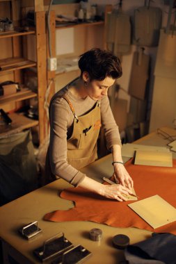 Atölyede çalışan bir kadın dikkatle deriyi kesiyor ve şekillendiriyor. Etrafı alet ve malzemelerle çevrili. El yapımı deri ürünler oluşturma sürecine girdi