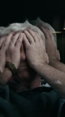 Beyaz saçlı, yaşlı beyaz bir adamın dikey pozu. Yüzüne sürtünüyor. Karanlık oturma odasında yalnız başına kalmaktan huzursuz ve kafası karışık.