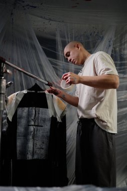 Side view of male artist with buzz cut applying adhesive tape to designer jacket creating exclusive design with grunge white print, while working in workshop all covered in masking film clipart