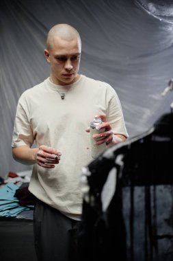 Orta boy erkek moda sanatçısı elinde beyaz boyalı eski ceketle resim stüdyosunda çalışırken elinde aerosol kutusu tutarken.