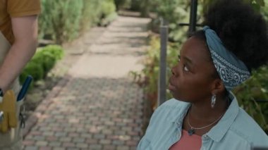 Genç Afrikalı Amerikalı bir kadının el kamerasıyla çekilmiş fotoğrafı. Açık hava bitkilerinde dizüstü bilgisayarda çalışırken. Beyaz tenli bir bahçıvanın parlak yaz gününde el arabasında taşıdığı çanak çömlek fidelerini kontrol ediyor.