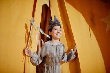 Smiling child wearing a crown and holding a magic wand, standing in a theater-like environment with yellow curtains, bringing light and joy to the scene through imagination and innocence clipart