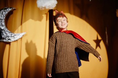 Kahverengi süveter ve kırmızı pelerin giymiş bir çocuk hilal pervaneli ve sarı arka planda gülümseyen bir sahnede duruyor. Arkaplandaki gölge sanatsal bir etki yaratıyor