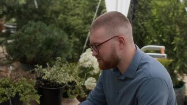 Gündelik giyimli ve gözlüklü genç beyaz adam yazın açık bahçe köşkündeki masada oturuyor, bilgisayarında konteynırdaki saksılardan birini gösteriyor.