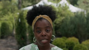 Kendine güvenen, halka küpeli genç Afro-Amerikan kadının yakın çekimi. Kameraya konuşan, yemyeşil yaz bahçesinde, bulanık arka planda beyaz bir köşkü olan.
