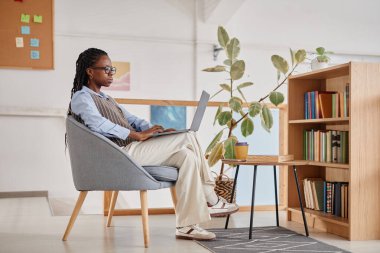 Arka planda bitki ve kitaplık bulunan rahat bir ev ofisinde çalışmaya yoğunlaşan kişi, verimli ve rahat bir çalışma alanı yaratıyor
