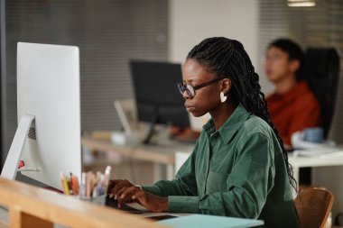 Masada oturan bir kadın arka planda meslektaşıyla bilgisayarla çalışıyor. Ofis alanı modern teknoloji ve organize çalışma alanı ile donatılmış verimli bir ortam yaratıyor